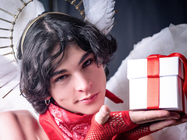Close-up de LukeHowlandy com cabelo preto encaracolado, asas de anjo e um halo, segurando uma caixa de presente vermelha e branca. Usando luvas vermelhas de renda, olhando diretamente para a câmera com um sorriso discreto..