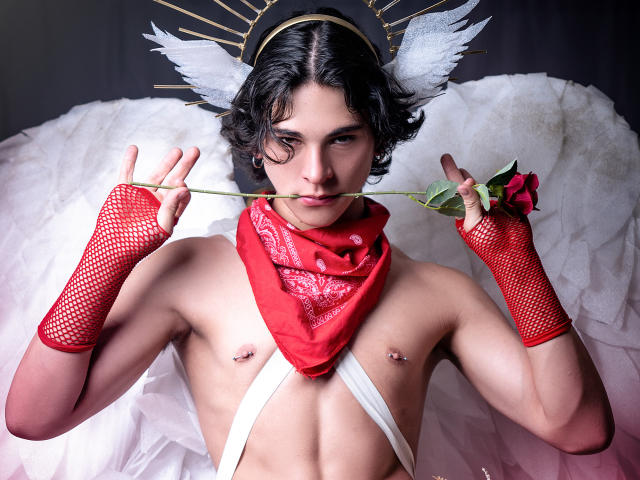 Luke Howlandy, com cabelo preto, usando um lenço vermelho, asas brancas, luvas de malha vermelhas e uma faixa branca enrolada no peito, segurando uma rosa entre os dentes, com um olhar sedutor.