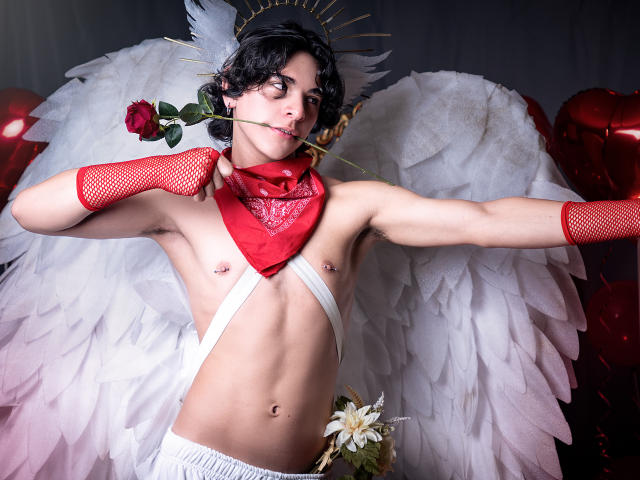 LukeHowlandy, torse nu, ailes d'ange blanches, gants en filet rouges, bandana rouge, rose dans la bouche, tissu blanc autour de la taille, fleur dans l'entrebaine, cheveux noirs et bouclés, éclairage tamisé, érotique, regard intense, pose suggestive..