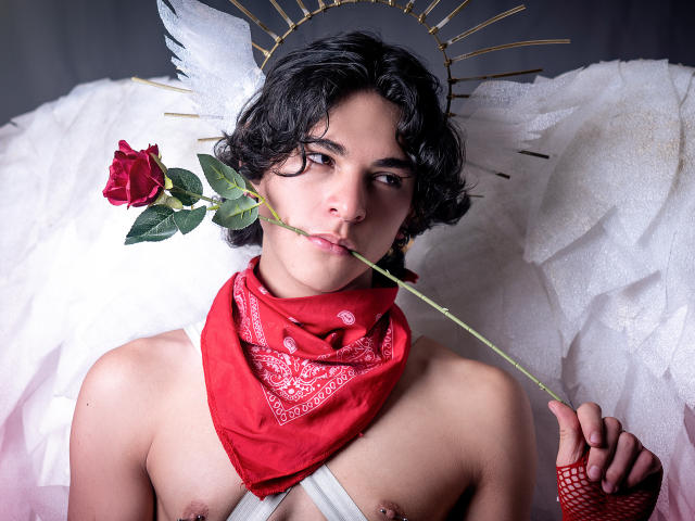 Luke Howlandy, sin camiseta, con cabello negro rizado, sosteniendo un tallo de rosa en la boca, con un pañuelo rojo alrededor del cuello, alas blancas de ángel, halo dorado, piercings en los pezones, tensión sexual, ojos oscuros, mirada seductora.