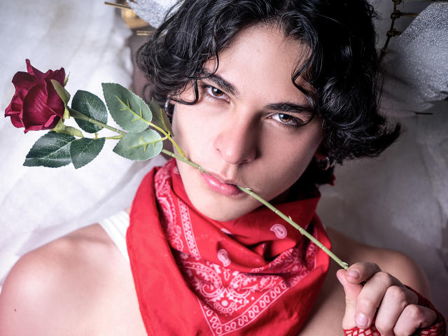 Luke Howlandy, com cabelos pretos encaracolados, segura uma rosa vermelha perto dos lábios, usando um lenço vermelho na cabeça. Eles têm olhos intensos e uma expressão ligeiramente provocadora. Uma rosa vermelha vibrante contrasta com a pele clara..