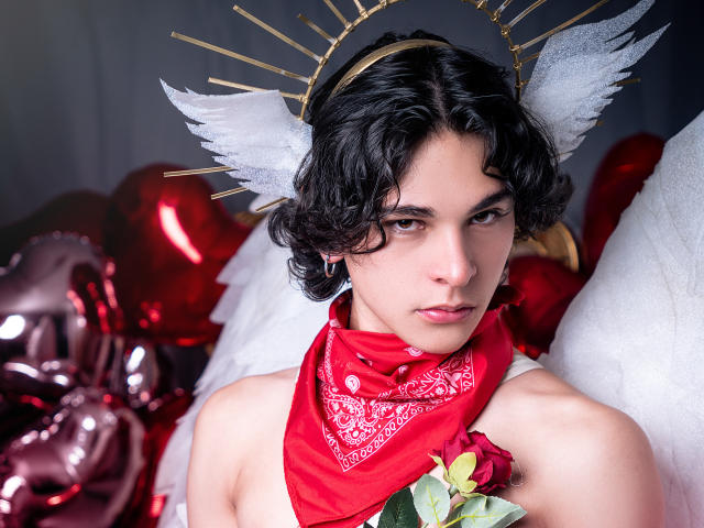 Luke Howlandy mit kurzen, lockigen schwarzen Haaren, der einen goldenen Heiligenschein mit weißen Flügeln, ein rotes Bandana und eine Blumenbluse trägt. Dekoration, intensiver Blick, umgeben von glänzenden roten Ballons, erotische Spannung.