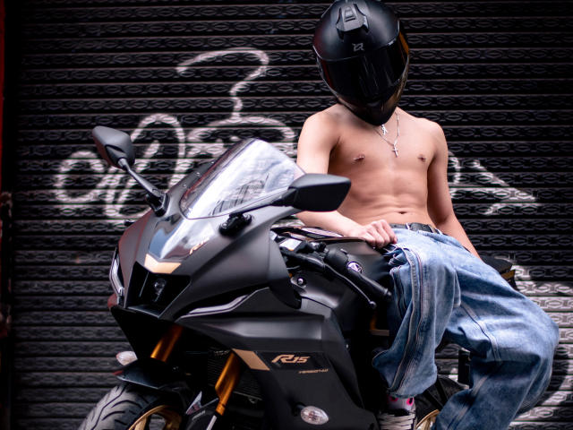 Luke Howlandy, sem camisa, usando um capacete preto e calças jeans largas, está sentado em uma motocicleta preta com faróis brilhantes. Grafite em um fundo de parede escura. Tensão sexual evidente devido ao tronco exposto e à pose casual..