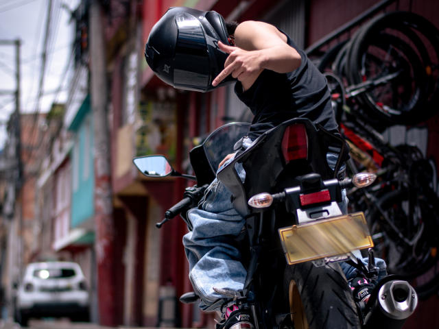 Luke Howlandy, vestindo uma jaqueta de couro preta e capacete, apontando com a mão direita, apoiado em uma motocicleta preta com detalhes cromados. Detalhes em um beco movimentado na área urbana. Cena de rua desfocada ao fundo.