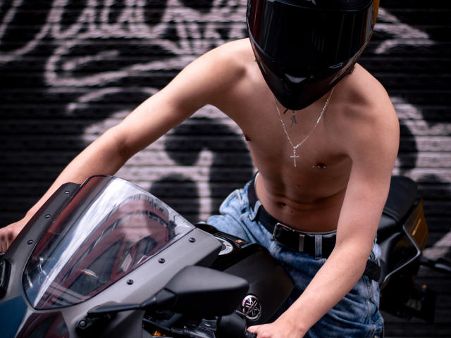 Luke Howlandy sem camisa, usando um capacete preto, pilotando uma motocicleta preta com um para-brisa transparente. Vestindo calças jeans azuis, um cinto e um colar com uma cruz de prata. Fundo com grafite preto. Musculoso, tenso, sexy.