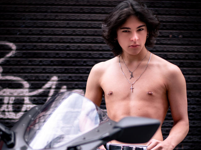 Luke Howlandy, sem camisa, com cabelo preto ondulado e porte médio, usando um colar com uma cruz, está em frente a uma parede de tijolos pretos. Parede com um farol de motocicleta brilhante em primeiro plano. Muito atraente.