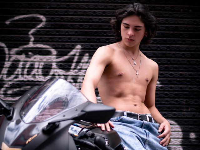 Luke Howlandy, sem camisa, com cabelo preto curto, vestindo calças jeans de cintura baixa, está encostado em uma motocicleta preta, olhando para baixo. O fundo é uma parede de tijolos coberta por grafites. Os mamilos de Luke estão visíveis, criando uma atmosfera sensual..