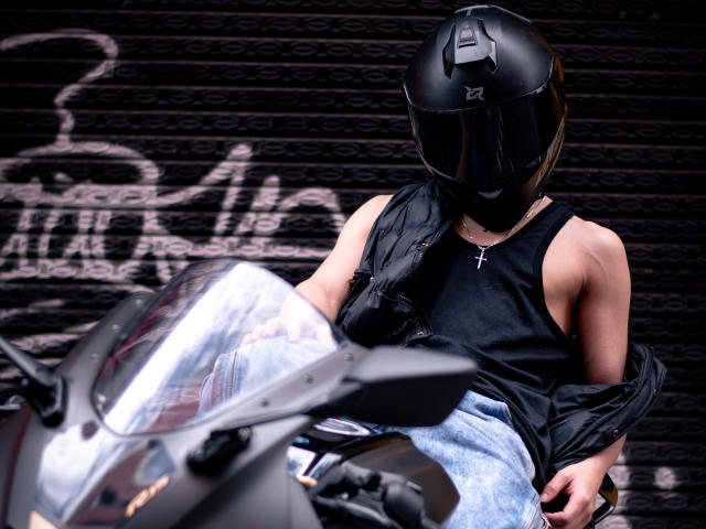 Digital artwork of LukeHowlandy in a black helmet, sleeveless black shirt, and blue jeans, sitting on a motorcycle with bright headlights. Background has blurred graffiti text. Sexual tension.