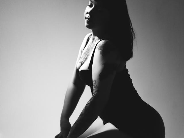 Photographie en noir et blanc d'AlinaMatei en profil, agenouillée, le dos tourné vers l'appareil photo. Elle porte un haut ajusté en dentelle qui met en valeur ses courbes et sa poitrine généreuse. L'éclairage à contraste élevé crée des ombres saisissantes..