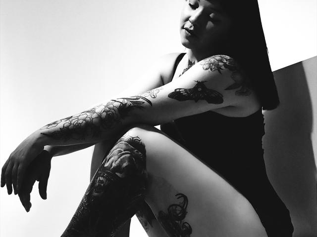 Fotografía en blanco y negro de Alina Matei sentada con las piernas cruzadas y los brazos apoyados en las rodillas, mostrando tatuajes detallados. en los brazos y los muslos. Alto contraste, sombrío, tensión erótica, fondo blanco puro..