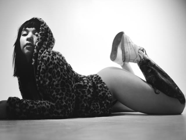 Fotografía en blanco y negro de AlinaMatei acostada boca abajo, con las piernas dobladas a las rodillas, vistiendo un suéter con estampado de leopardo. mostrando sus muslos desnudos y zapatillas blancas, mirada intensa, iluminación de alto contraste..