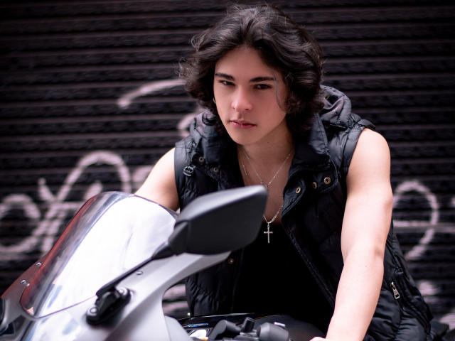 Luke Howlandy, con el pelo negro despeinado, se inclina hacia adelante sobre una motocicleta blanca, vistiendo un chaleco negro y un crucifijo. Con un aspecto intenso, fondo negro con textura y grafitis blancos. Tensión sexual, mirada fija..