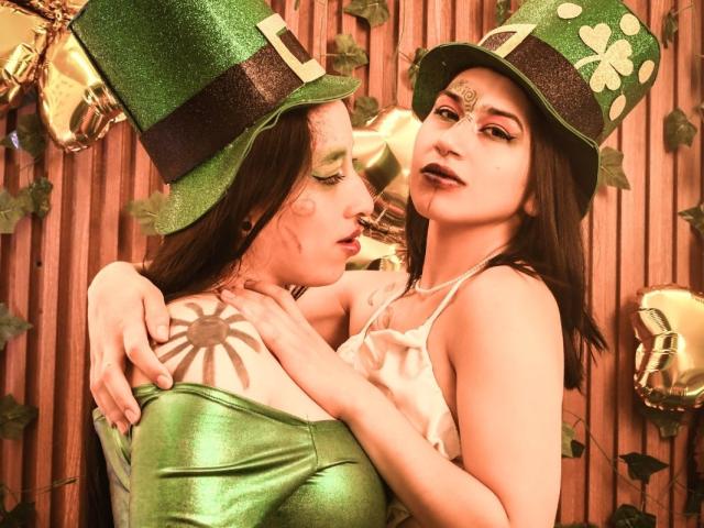 Duas mulheres da HotHarmonyX, usando chapéus verdes com glitter e detalhes de trevo, uma vestindo um vestido verde e a outra um vestido branco. Abraçando, olhos semicerrados, tensão sexual, fundo de madeira clara..