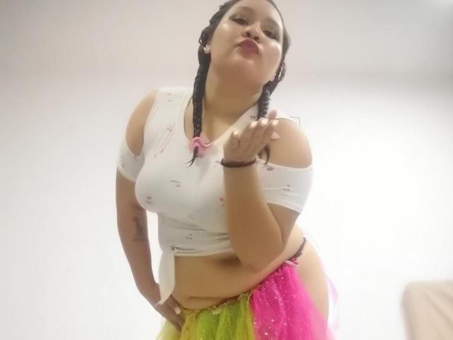 CattaleyaBohorquez stands low-angle, looking up with a seductive expression. She wears a white off-shoulder top, revealing her belly, and a neon pink and yellow skirt. Her hand rests on her crotch.