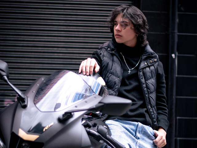 Luke Howlandy está sentado em uma scooter preta, vestindo uma jaqueta puffer preta e calças jeans azuis. Ele se apoia no guidão, olhando atentamente. Faróis brilhantes em primeiro plano. Tensão sexual evidente..