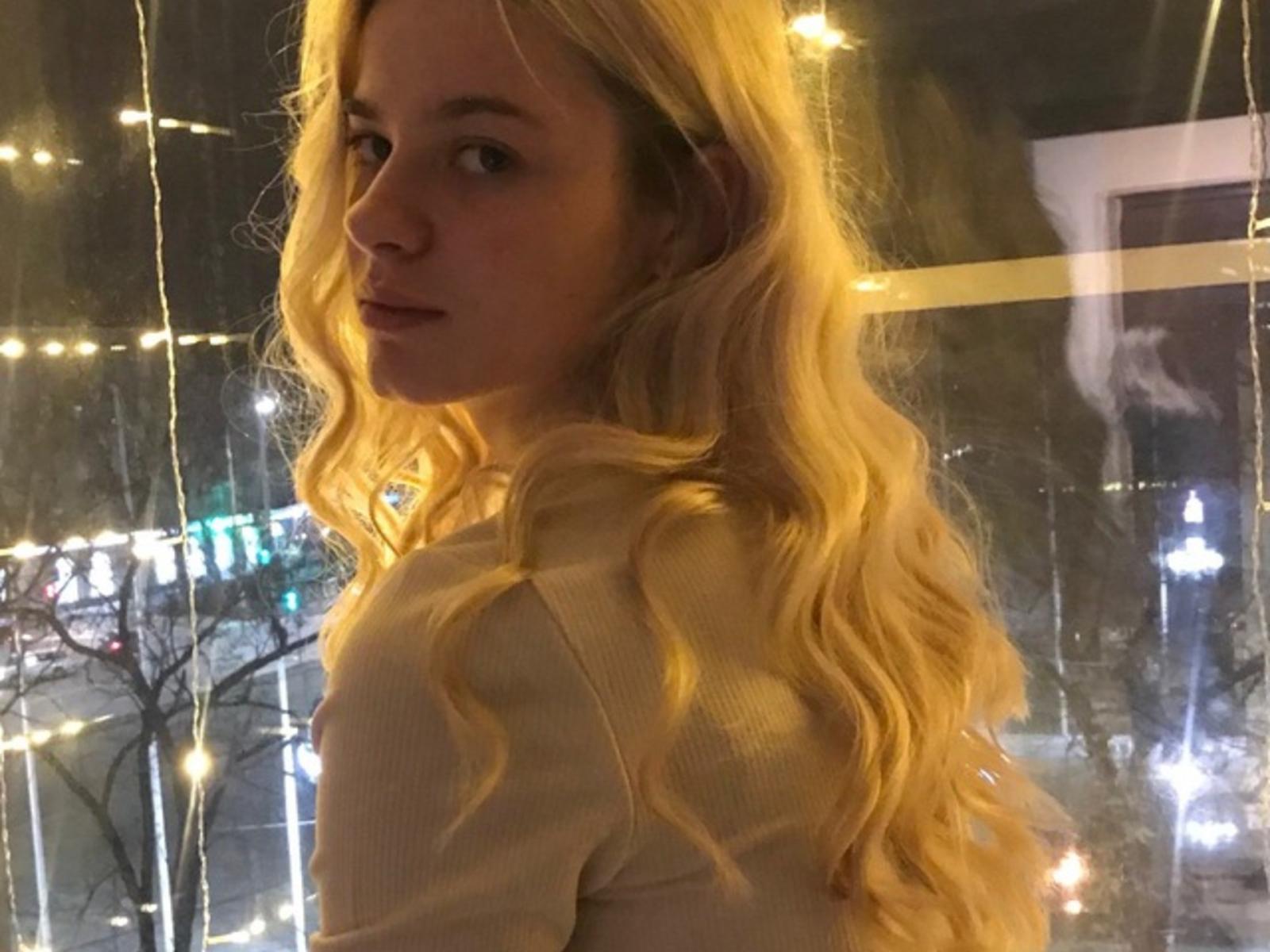 Fotografie einer blonden Frau mit welligem Haar, die über ihre Schulter schaut, beleuchtet von leuchtenden Lichterketten. Sie trägt ein beiges Oberteil, ihr Ausdruck ist sinnlich und erzeugt sexuelle Spannung..