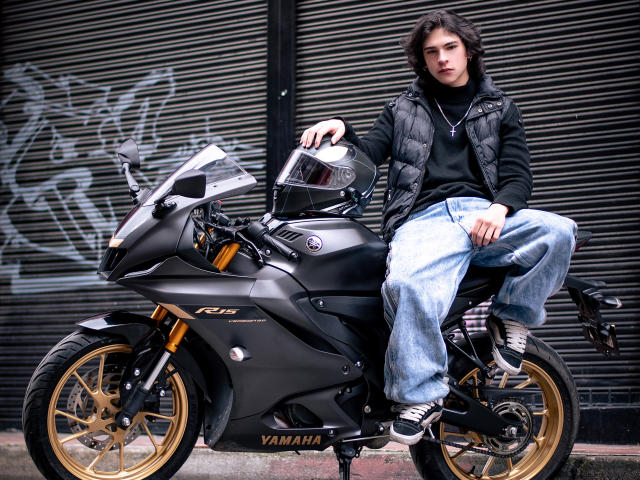 Luke Howlandy está sentado em uma moto esportiva preta da Yamaha, com rodas douradas, vestindo uma jaqueta preta acolchoada e calças jeans azuis largas. Inclinado de forma relaxada, com um cigarro na mão. Fundo com grafite urbano.