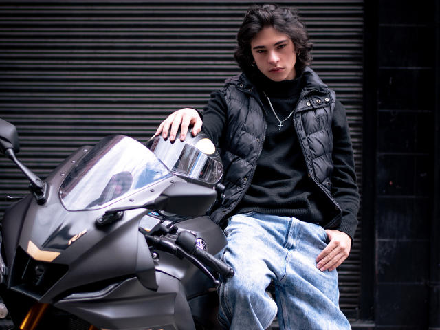 Luke Howlandy, met krullend zwart haar, gekleed in een zwarte gewatteerde jas en blauwe jeans, leunt tegen een zwarte motorfiets. Wit vizier, met een serieuze blik. Donkere, gestructureerde achtergrond. Seksuele spanning..