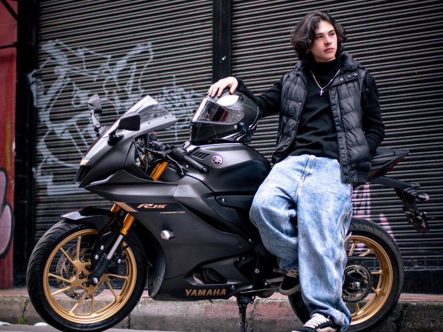 Luke Howlandy seduto su una motocicletta Yamaha nera con cerchi dorati, indossa una giacca imbottita nera e jeans blu larghi. Mano sulla visiera, sfondo urbano con graffiti. Posizione incredibilmente sexy..