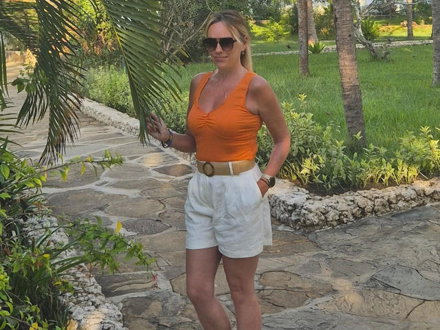 Sofie, vestida con una camiseta naranja y pantalones cortos blancos, está de pie al aire libre en un camino de piedra. Ella lleva gafas de sol y sostiene una hoja de palma. Su cuerpo está en forma, con un escote visible..