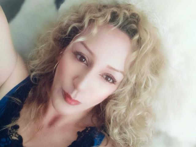 Autoportrait en gros plan de SammyDesire, avec des cheveux blonds bouclés, du rouge à lèvres rouge et un eye-liner foncé, portant un haut en dentelle noire. regardant directement l'appareil photo avec une expression légèrement provocante.