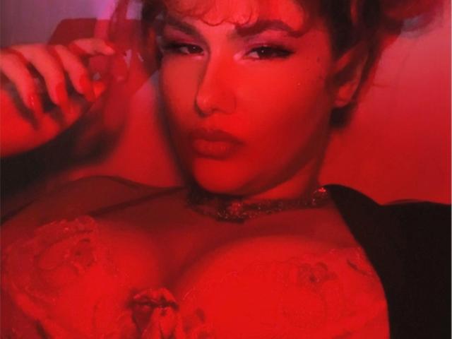 Close-up van DelirioPerpetuo, een aantrekkelijke vrouw met rood licht, die een telefoon op haar oor houdt. Ze heeft grote borsten in een rode kanten beha, en ze ziet er verleidelijk uit. Een rode gloed accentueert haar rondingen..