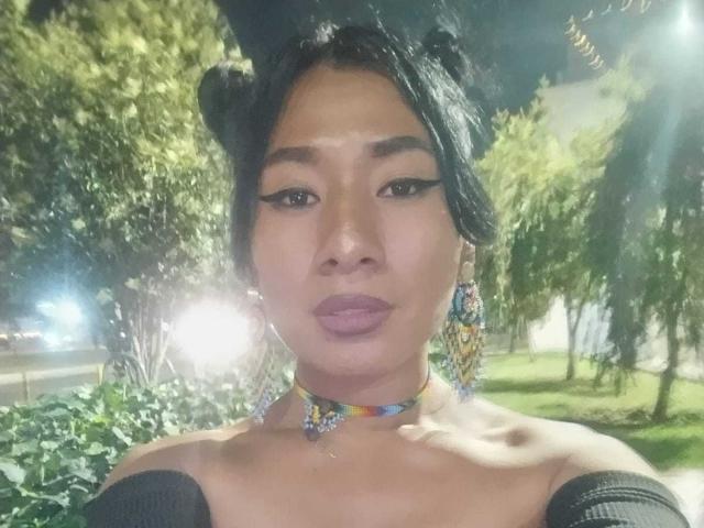 Close-up of JatziBehot69, a woman with dark hair in two buns, wearing a colorful choker and dangling earrings, with full lips and dark eyeshadow, in a sunlit park. Flirty expression.