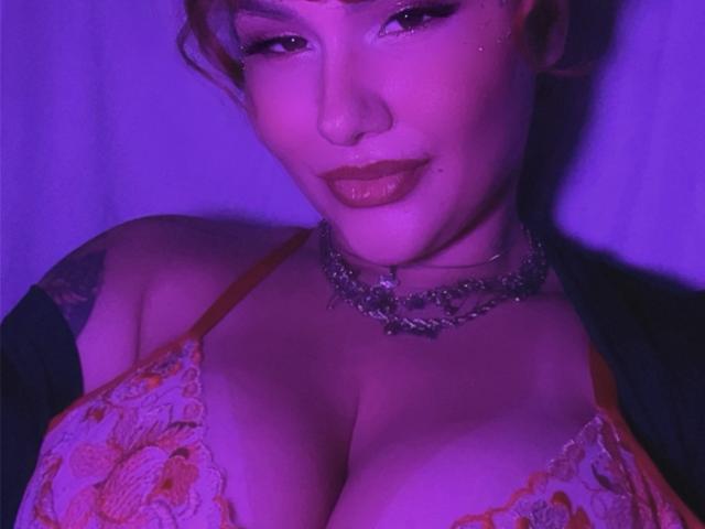 Close-up photo of DelirioPerpetuo, face partially illuminated by purple light, wearing a floral lace bra that accentuates large breasts. Choker necklace around neck, slightly seductive expression, lips slightly parted.