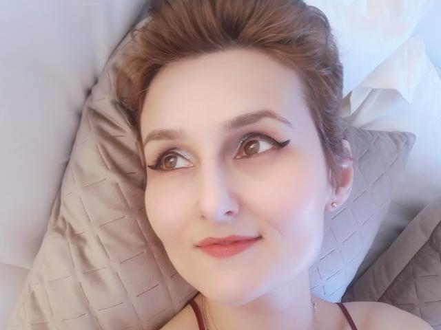 Fotografía de primer plano de LamourCestMoi, una mujer de piel clara y cabello castaño, acostada sobre un cojín acolchado. Ella tiene ojos marrones, delineador de ojos negro y lápiz labial rojo, y mira hacia arriba con una ligera sonrisa. Su blusa muestra un pequeño detalle de una correa..