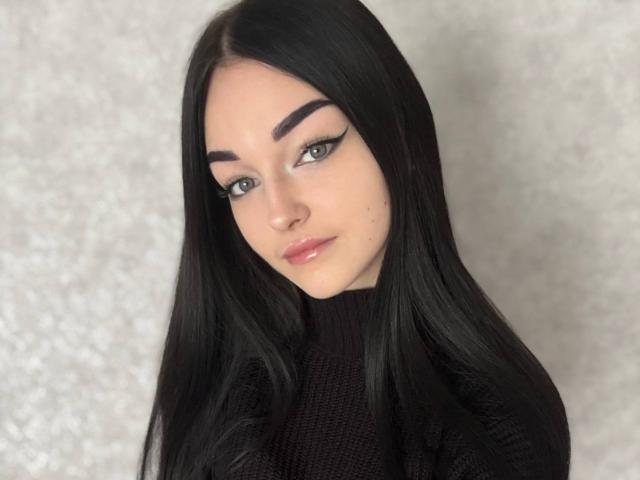 Roseana Conlin mit langem, schwarzem Haar, heller Haut und grünen Augen, trägt einen schwarzen Rollkragenpullover. Sie blickt direkt in die Kamera, mit einem leichten Lächeln, und wirkt dabei sexy und selbstbewusst..