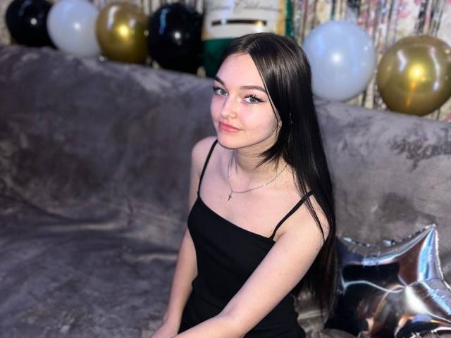 RoseanaConlin with long black hair, wearing a black spaghetti strap dress, sitting on a gray velvet couch, surrounded by gold and white balloons, looking directly at the camera, slightly smiling.