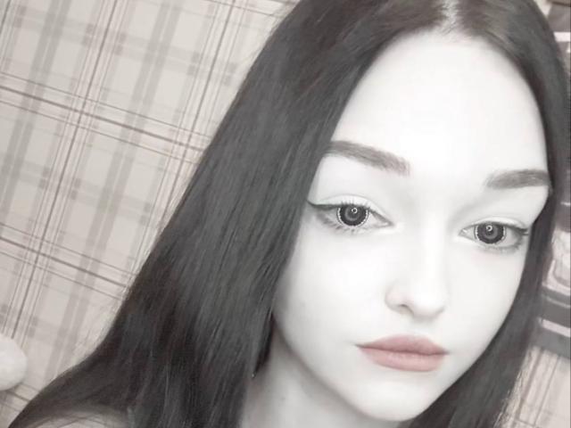 Photographie en gros plan de RoseanaConlin, visage principalement blanc avec des cheveux noirs, fond à carreaux, maquillage prononcé, partiellement effacé. visage, regard intense, léger sourire, yeux surexposés, lèvres roses et douces, tension sexuelle, ambiance étrange..