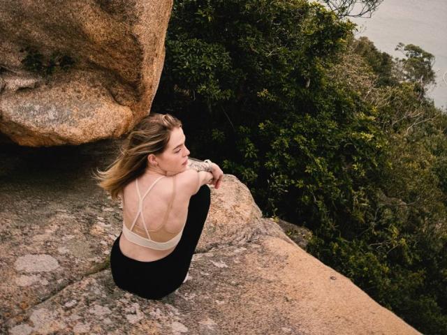 MaryanBatteen est accroupie sur une corniche rocheuse, le dos tourné vers la caméra, et porte un soutien-gorge beige et un pantalon noir. Elle regarde à droite, les mains posées sur ses genoux, avec une végétation luxuriante derrière elle. Extrêmement séduisant..
