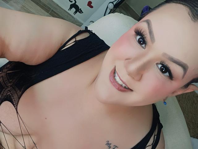 Close-up selfie of GiiannaMorrys, smiling with pink lips, black mesh top revealing cleavage, nose ring, blue earrings, light skin, dark brown hair, tattoo on shoulder, cock visible in background.