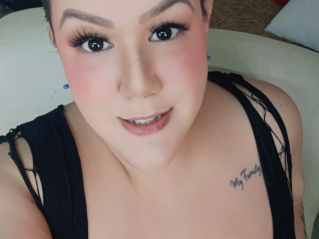 Foto em close-up de GiiannaMorrys, com pele clara, sobrancelhas escuras e delineador preto. Ela tem um piercing no nariz, uma camiseta preta com alças desfiadas e uma tatuagem no ombro direito. Muito fofo.