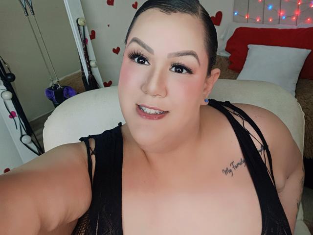 GiiannaMorrys, a curvy woman with fair skin and dark hair, wears a black lace top, smiling at the camera. She has a "My Love" tattoo on her right shoulder. Red heart decorations and string lights in the background.