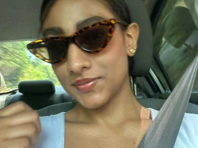 Close-up selfie of NatalyBlonder in a car, wearing dark tortoiseshell sunglasses, white top, and gold hoop earrings, holding seatbelt, with green blurry background, slightly smiling, natural light.