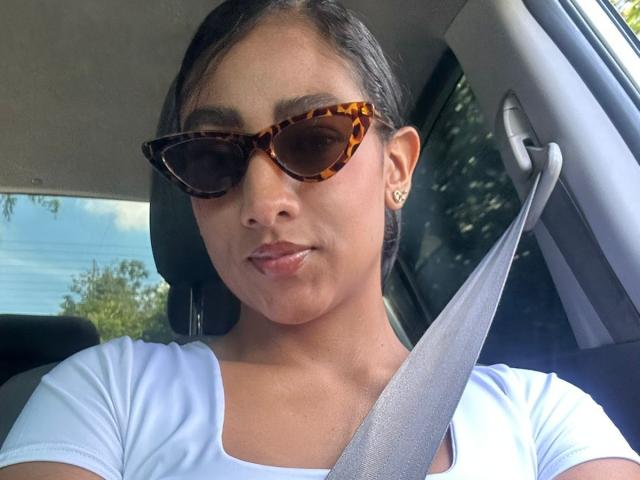 NatalyBlonder with tan skin and dark hair, wearing tortoiseshell sunglasses and a white t-shirt, sitting in a car with a seatbelt across her chest, looking directly at the camera.