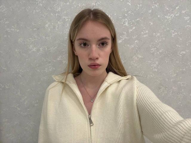 CelsaHoeger a les cheveux châtain clair, et elle porte un pull zippé de couleur crème, sur un fond gris texturé. Elle regarde directement l'appareil photo avec une expression neutre, une légère rougeur sur ses joues..