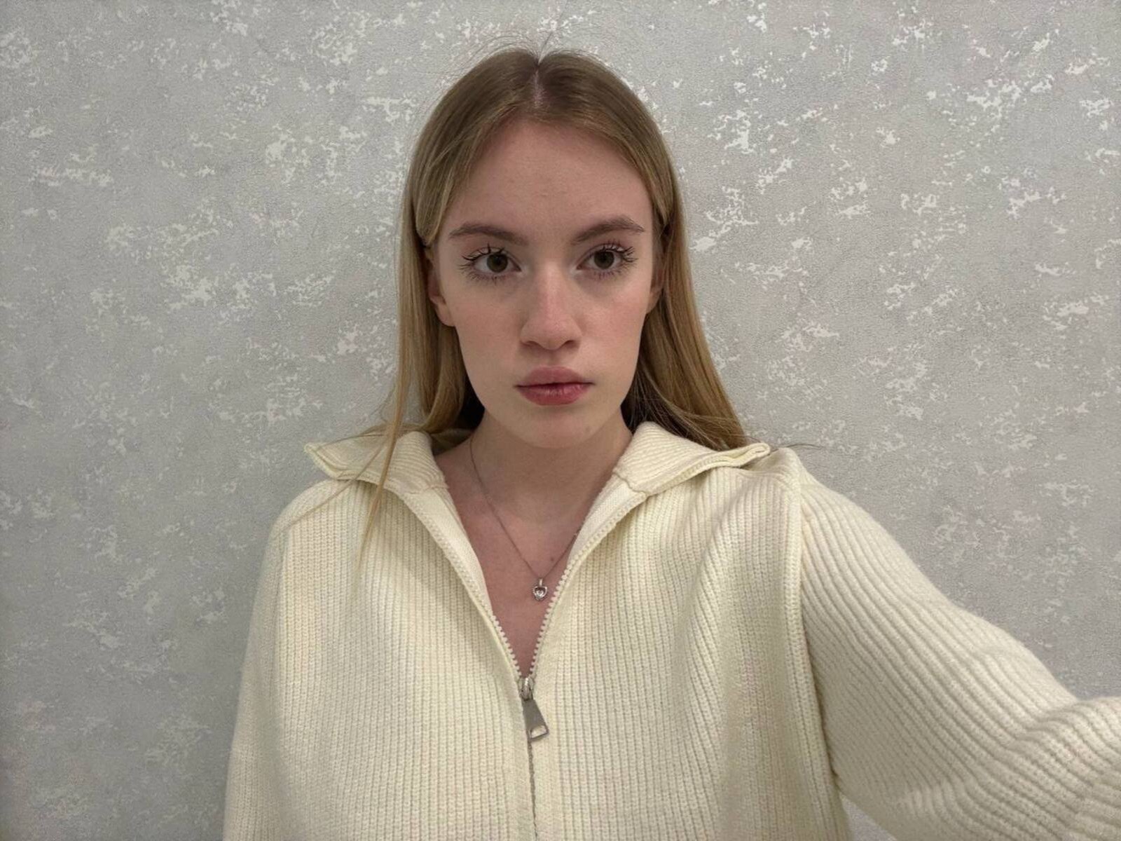 CelsaHoeger ha i capelli castano chiaro, indossa un maglione beige con zip, su uno sfondo grigio con una texture. Guarda direttamente nella fotocamera con un'espressione neutra, con un leggero rossore sulle guance..