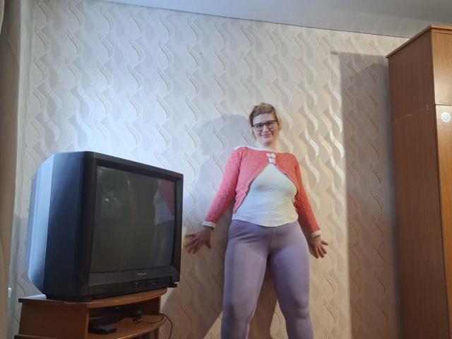 SabrinaShine está em um quarto com papel de parede texturizado, vestindo uma blusa vermelha de manga comprida e uma camiseta branca, e calças cinzas justas. calças. Uma televisão antiga em um suporte à sua esquerda. Sombras projetadas na parede..