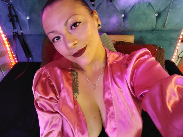 Photograph of SusanaHotSex, close-up, wearing a shiny pink satin robe, deep cleavage, tattoo on left chest, dark lipstick, black earrings, blue-tufted headboard, neon lights in background, sexy, seductive pose.