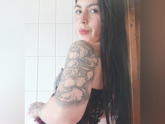 Fotografia di EriiMiilk, che guarda parzialmente di lato, con la testa girata, capelli neri, braccio tatuato, top di pizzo nero, chiaro. pelle, luce naturale, sorriso dolce, sfondo di piastrelle bianche, cornice in legno, tensione sessuale.