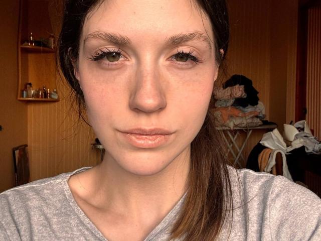 Fotografia ravvicinata di Rayna Niffenegger, con la pelle chiara, occhi marroni, un trucco leggero e capelli castani lisci. Indossa una camicia grigia e guarda direttamente la fotocamera con un'espressione neutra. Lo sfondo mostra una stanza disordinata..