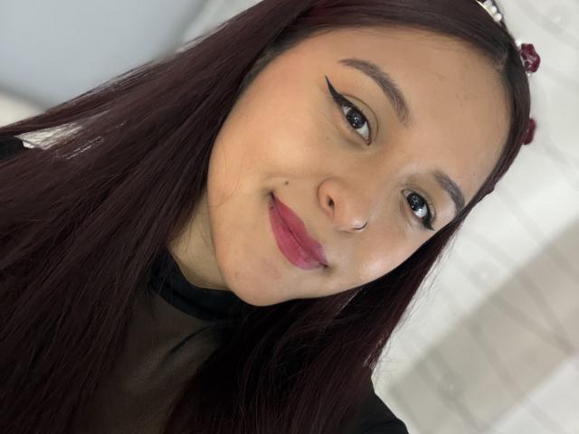 Close-up photo of MarianaVargasX with long, straight black hair, wearing a black mesh top. She has a nose piercing, winged eyeliner, and pink lipstick. Background is blurred white. Fucking cute.