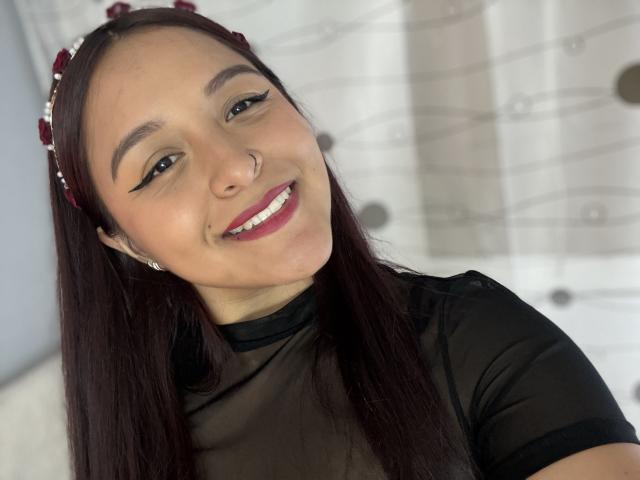 Primer plano de MarianaVargasX con cabello negro largo, una diadema roja, una blusa negra transparente, sonriendo con lápiz labial rojo, y mostrando su nariz. piercing y delineador de ojos negro. Luz suave en el fondo. Increíblemente atractivo..