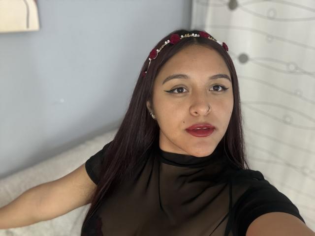 Nahaufnahme von MarianaVargasX, einer lateinamerikanischen Frau mit langen schwarzen Haaren, rotem Lippenstift und einem schwarzen, transparenten Oberteil. Sie blickt mit einem leichten Lächeln in die Kamera und zeigt dabei einen Hauch von Ausschnitt. Heller Raumhintergrund.