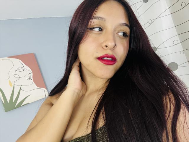 Close-up photo of MarianaVargasX, a Latina woman with long black hair, red lipstick, and a hand touching her neck. She's looking to the side with a slightly aroused expression.