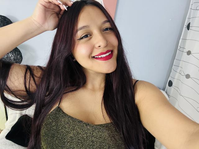 Fotografia de MarianaVargasX com cabelo preto longo e liso, batom vermelho e um piercing no nariz. Ela está sorrindo, vestindo uma blusa verde brilhante, com a mão no cabelo, e o braço direito levantado. Tensão sexual.