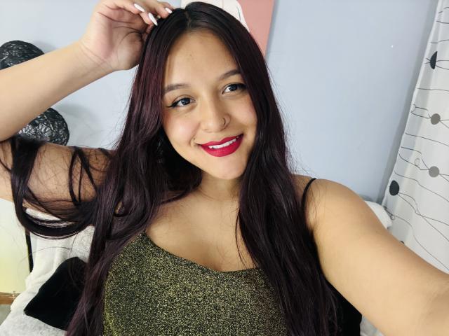 Photographie de MarianaVargasX avec de longs cheveux noirs et lisses, du rouge à lèvres rouge et un haut vert scintillant, souriant directement. devant l'appareil photo.
elle touche sa tête avec une main, dégageant une tension sexuelle..